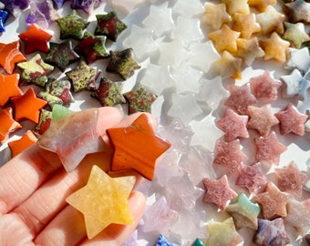May include: A collection of star-shaped polished stones in various colours and patterns, including red, orange, yellow, green, blue, purple, and white. The stones are arranged on a white surface.