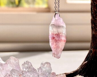 May include: A pink and white crystal pendant on a silver chain, hanging above a cluster of pink and white crystals.