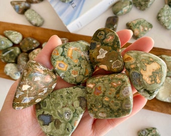 May include: A collection of green, brown, and white tumbled stones with a speckled pattern. The stones are smooth and polished, and they are arranged in a hand.