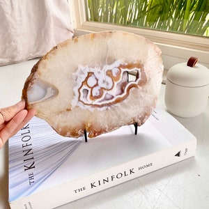 May include: A large, white agate slice with brown and orange banding. The agate is on a black metal stand and is displayed on a white book with the text "The Kinfolk Home".