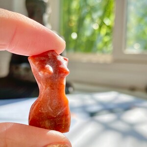 May include: A small, hand-carved, red-brown stone sculpture of a female torso. The sculpture is smooth and polished, with a slightly textured surface.