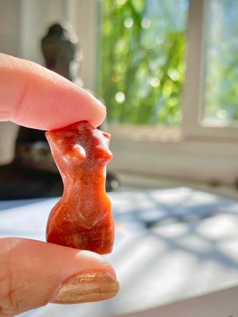 May include: A small, hand-carved, red-brown stone sculpture of a female torso. The sculpture is smooth and polished, with a slightly textured surface.