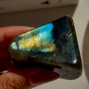 May include: A polished labradorite gemstone with a vibrant display of color. The stone features a mix of blues, yellows, and greens, with dark gray and black undertones. The surface is smooth and reflective, showcasing the stone's natural beauty.