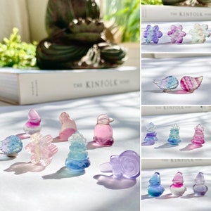 May include: A collection of colorful, iridescent glass figurines in various shapes, including a bird, a tree, a snowflake, a snail, and a butterfly. The figurines are arranged on a white surface with a soft, natural light.