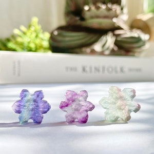 May include: Three iridescent snowflake-shaped fluorite crystals in shades of purple, green, and blue. The crystals are arranged in a row on a white surface.