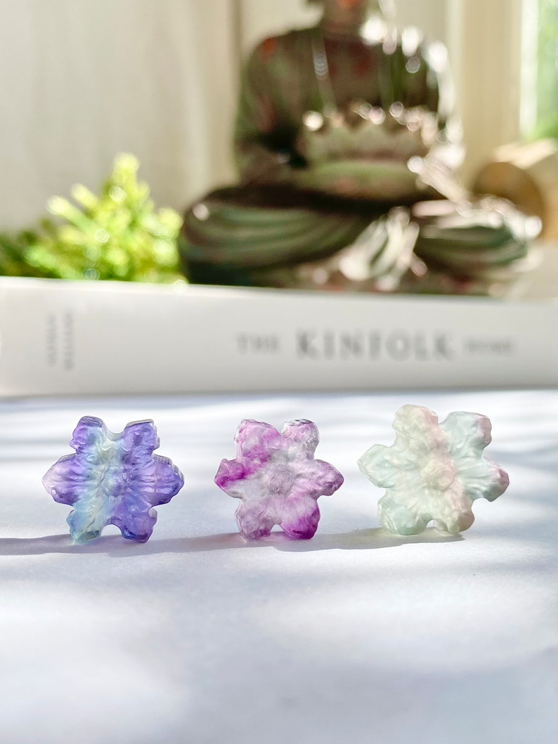 May include: Three iridescent snowflake-shaped fluorite crystals in shades of purple, green, and blue. The crystals are arranged in a row on a white surface.