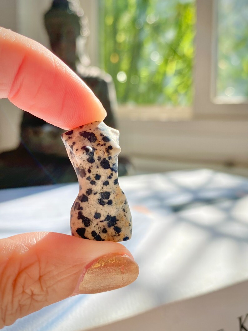May include: A small, hand-carved, dalmatian jasper figurine of a female torso. The figurine is a light beige colour with black spots.