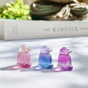 May include: Three small glass penguin figurines, each with a different color gradient. The penguin on the left is pink and white, the middle penguin is blue and white, and the penguin on the right is purple and white.