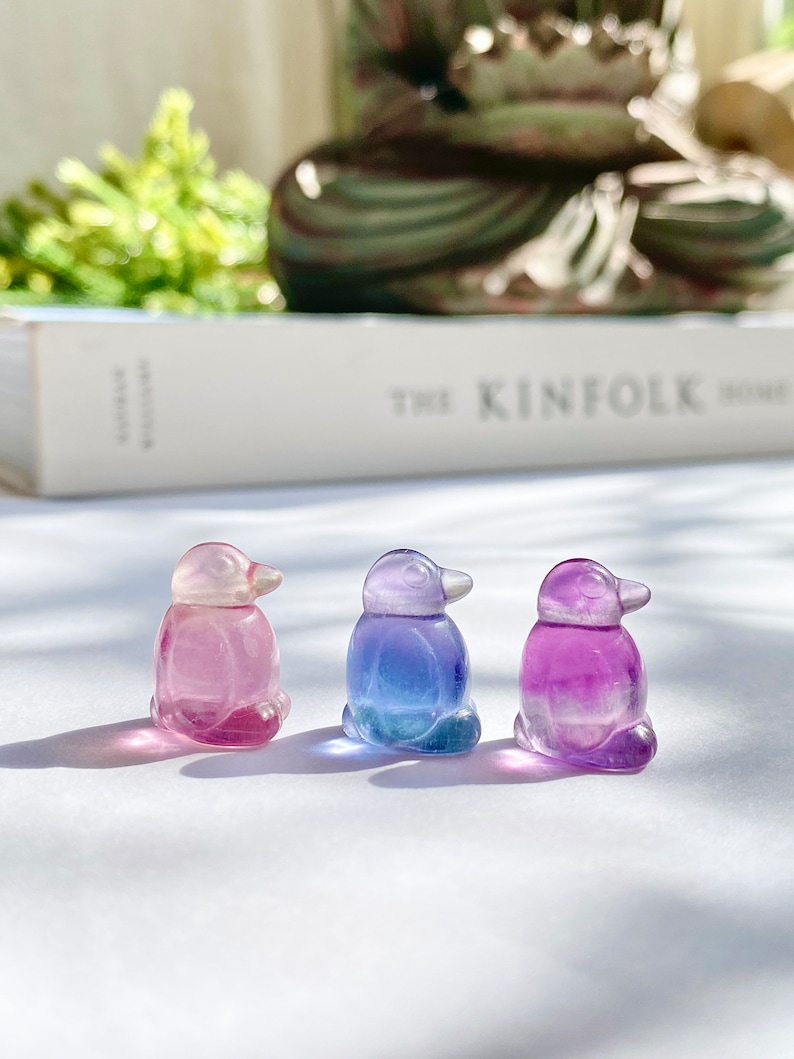 May include: Three small glass penguin figurines, each with a different color gradient. The penguin on the left is pink and white, the middle penguin is blue and white, and the penguin on the right is purple and white.