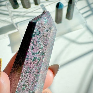 May include: A purple and green crystal point with a speckled pattern. The crystal is pointed at the top and has a flat base.