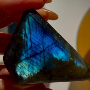May include: A triangular-shaped labradorite gemstone with a striking display of blue iridescence. The stone's surface showcases a play of light, with flashes of blue and hints of green. The gemstone is held in a hand, highlighting its natural beauty and unique color.