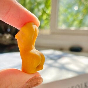 May include: A small, yellow, ceramic sculpture of a human figure. The sculpture is being held by a finger and thumb.