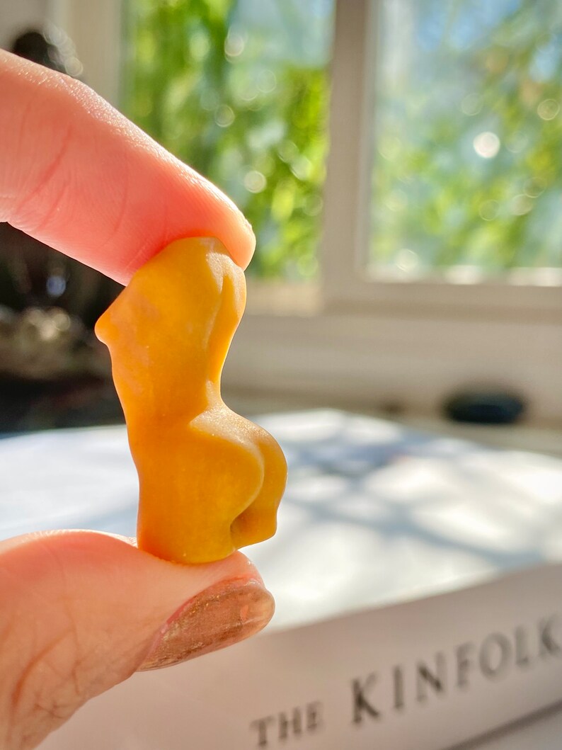 May include: A small, yellow, ceramic sculpture of a human figure. The sculpture is being held by a finger and thumb.