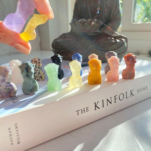 May include: A collection of 12 small, hand-carved, colourful stone figurines in various shapes and colours, arranged in a row on a white book with the text "THE KINFOLK HOME" on the cover.