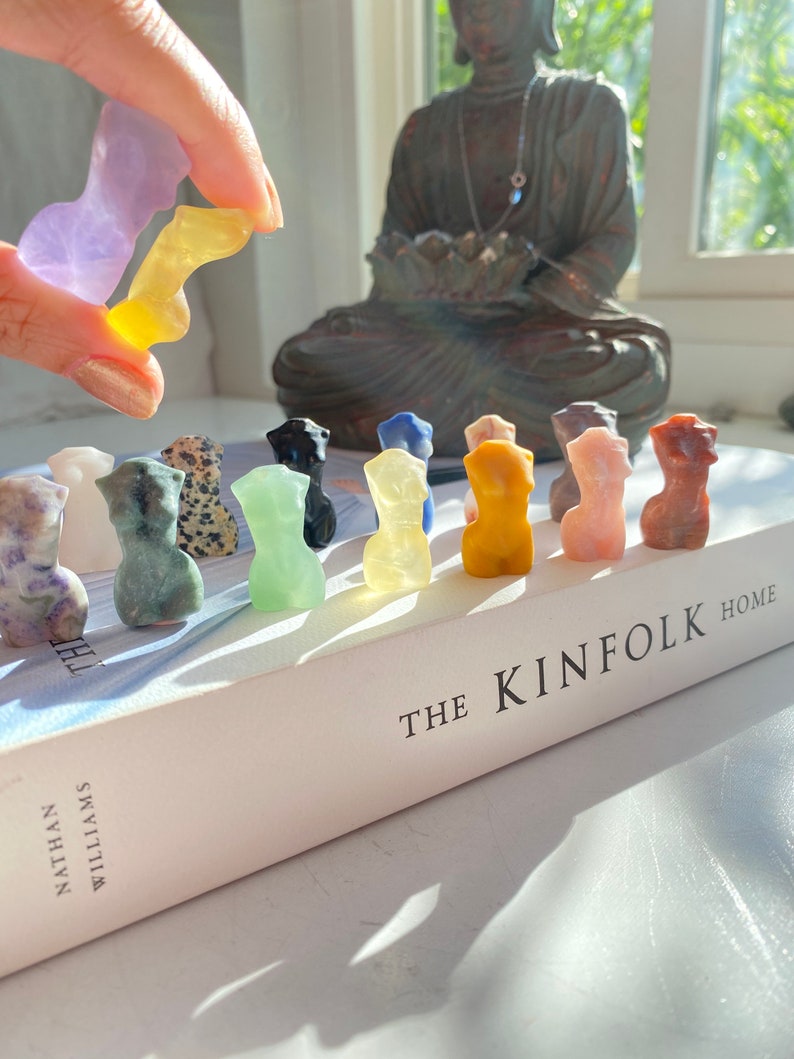 May include: A collection of 12 small, hand-carved, colourful stone figurines in various shapes and colours, arranged in a row on a white book with the text "THE KINFOLK HOME" on the cover.