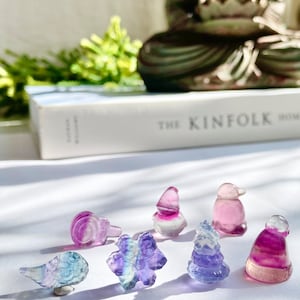 May include: A collection of seven small, colorful, crystal figurines in the shapes of a bird, a flower, a tree, a hat, a penguin, and a gnome. The figurines are made of fluorite, a type of crystal known for its iridescent colors.