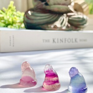 May include: Three small, hand-carved figurines in the shape of a person sitting with their head bowed. The figurines are made of a translucent, multi-colored material that appears to be fluorite. The figurines are arranged in a row on a white surface.