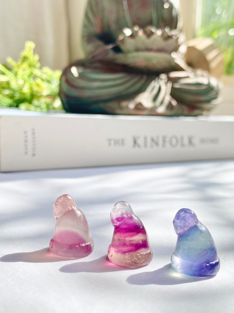 May include: Three small, hand-carved figurines in the shape of a person sitting with their head bowed. The figurines are made of a translucent, multi-colored material that appears to be fluorite. The figurines are arranged in a row on a white surface.