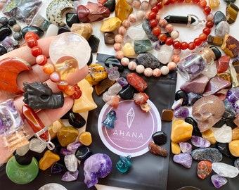 May include: A variety of crystals, stones, and beads in a variety of colors, including red, orange, yellow, green, blue, purple, and black. There are also several small glass bottles filled with crystals. The text "AHANA" is visible on a black bag.