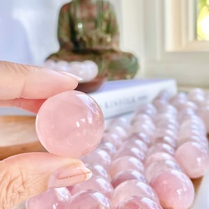 May include: A collection of pink rose quartz spheres, some of which are being held in a hand. The spheres are smooth and translucent, with a slight pink hue.