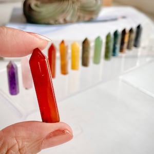 May include: A collection of polished, pointed crystal wands in various colors, including red, orange, yellow, green, blue, and purple. The wands are arranged in a row on a white surface.