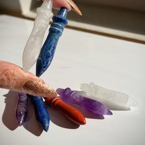 May include: A collection of gemstone wands in various colors, including clear quartz, lapis lazuli, red jasper, and amethyst. Each wand is approximately 4 inches long and features a pointed tip and a rounded handle.