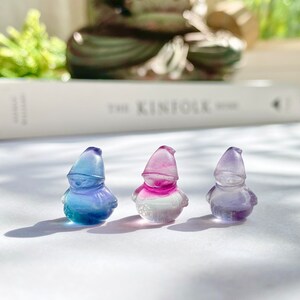 May include: Three small glass figurines of gnomes, each with a different color gradient. The gnome on the left is blue and green, the middle gnome is pink and white, and the gnome on the right is purple and white.