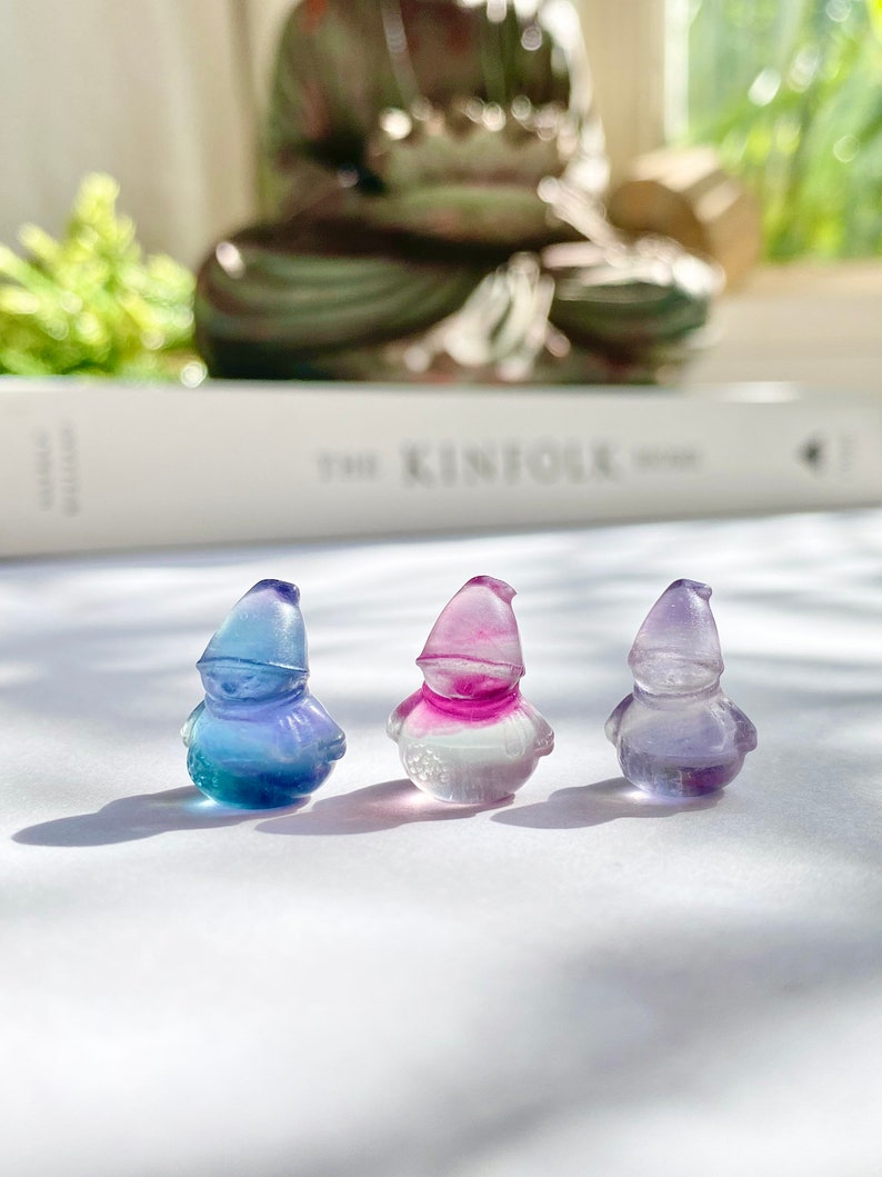 May include: Three small glass figurines of gnomes, each with a different color gradient. The gnome on the left is blue and green, the middle gnome is pink and white, and the gnome on the right is purple and white.
