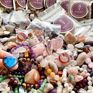 May include: A variety of colorful gemstones and crystals, including amethyst, rose quartz, tiger's eye, and labradorite, are displayed in a pile. Some of the stones are in small plastic bags labeled "Ahana".