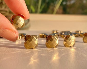 May include: Close-up of four gold-coloured metal buttons with a geometric design. The buttons are arranged in a row on a white surface.