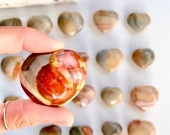 May include: A collection of heart-shaped stones in various shades of brown, beige, and red. One stone is held up, showcasing its polished surface. The background features more heart-shaped stones, suggesting a theme of love or natural decor.