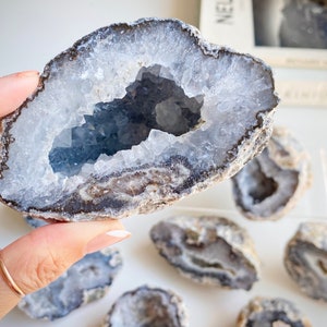 May include: A collection of agate geode crystals with a variety of sizes and shapes. The geodes are a light blue color with a white and gray outer layer. The crystals are arranged on a white surface.