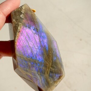 May include: A dark gray labradorite crystal with iridescent purple and blue flashes. The crystal is faceted and has a hexagonal shape.