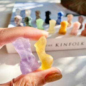 May include: Two small glass sculptures in the shape of the human form. One is a light purple colour and the other is a light yellow colour.