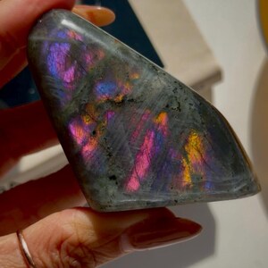 May include: A polished, triangular labradorite stone. The stone displays a dark gray base color with iridescent flashes of pink, purple, blue, and yellow. The gemstone is held in a hand, showcasing its unique color play.