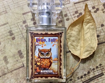 May include: A clear glass bottle of perfume with a gold label that says "Hello, Fall!" and features an illustration of an owl in a forest setting. The bottle is sitting on a sheet of music with a dried leaf.
