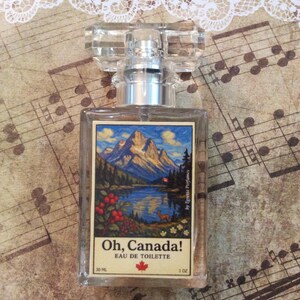 May include: A clear glass perfume bottle with a rectangular label featuring a mountain landscape scene. The label reads "Oh, Canada!" and "Eau de Toilette." The bottle has a silver collar and a clear, faceted cap. The bottle contains 1 oz of perfume.