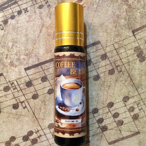May include: A perfume oil bottle with a gold cap and a label featuring a coffee cup illustration. The label reads "Coffee Beans" and "Perfume Oil". The bottle is dark with a gold cap.