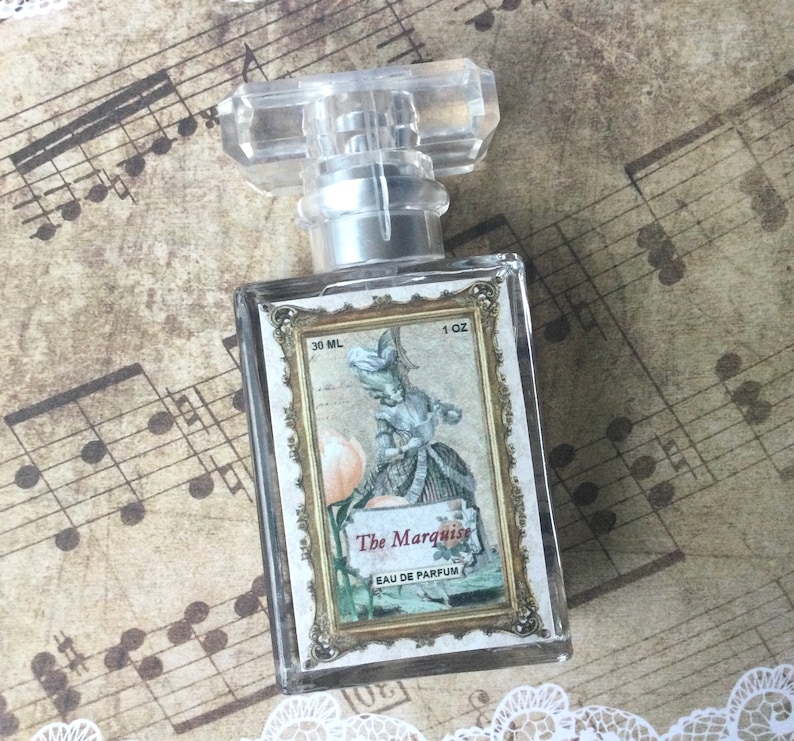 May include: A clear glass bottle of perfume with a gold cap and label. The label features a vintage illustration of a woman in a white dress and the text "The Marquise Eau de Parfum". The bottle is 30 ml or 1 oz.