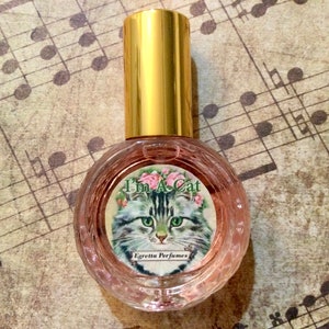 May include: A small, round glass bottle with a gold cap and a label that says "I'm A Cat" and "Egretia Perfumes". The label features a cartoon cat with green eyes and a pink rose.