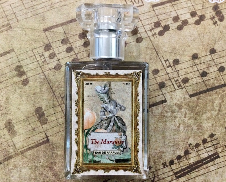 May include: A clear glass bottle of perfume with a gold-colored label. The label features a vintage illustration of a woman in a dress and the text "The Marquise Eau de Parfum". The bottle is 30 ml or 1 oz.