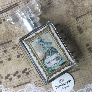 May include: A clear glass bottle of perfume with a gold-trimmed label that reads "The Marquise Eau de Parfum". The label features a vintage illustration of a woman in a dress holding a peach. The bottle is sitting on a sheet of music with a white circle sticker that reads "handmade for you".