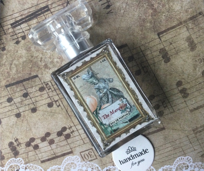 May include: A clear glass bottle of perfume with a gold-trimmed label that reads "The Marquise Eau de Parfum". The label features a vintage illustration of a woman in a dress holding a peach. The bottle is sitting on a sheet of music with a white circle sticker that reads "handmade for you".