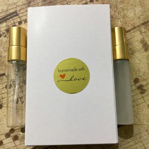 May include: A white box with a round sticker that reads "homemade with love" and a red heart. Two small perfume bottles with gold caps are next to the box. One bottle is clear, and the other is frosted.