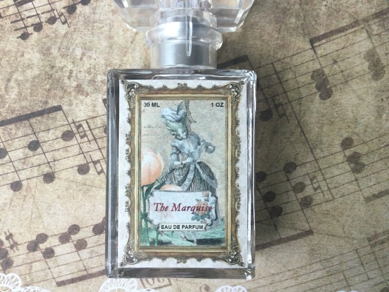 May include: A clear glass bottle of perfume with a gold-trimmed label. The label features a vintage illustration of a woman in a dress and the text "The Marquise Eau de Parfum 30 ML 1 OZ".