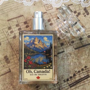 May include: A rectangular glass perfume bottle with a silver cap and a clear plastic cap. The label features a mountain landscape with the text "Oh, Canada!" and "Eau de Toilette". The bottle contains 1 oz of perfume.