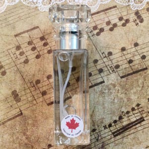May include: A clear glass perfume bottle with a faceted crystal stopper and silver-colored collar. The bottle has a white label with a red maple leaf and the text "Made in Canada / Fabriqu&eacute; au Canada". The background features sheet music.