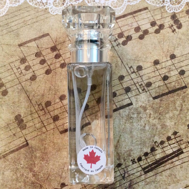 May include: A clear glass perfume bottle with a faceted crystal stopper and silver-colored collar. The bottle has a white label with a red maple leaf and the text "Made in Canada / Fabriqu&eacute; au Canada". The background features sheet music.