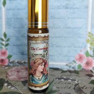 May include: A gold and brown glass roll-on bottle with a label that says "The Courtesan Perfume Oil". The label features a vintage illustration of a woman with a floral crown and a necklace.