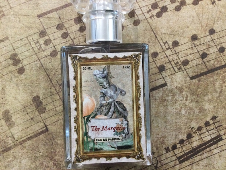 May include: A clear glass bottle of perfume with a gold-framed label. The label features a vintage illustration of a woman in a dress and the text "The Marquise Eau de Parfum 30 ML 1 OZ".